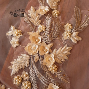 May include: Close-up of a floral embroidered applique with gold thread and faux pearls. The design features detailed leaves and flowers, with the text "Bridal Trove" in the upper left corner. The applique is on a sheer, blush-colored fabric.