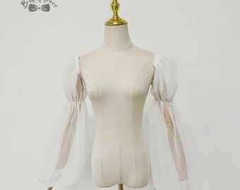 May include: A white, sheer bridal top with long, voluminous sleeves. The sleeves are gathered at the wrist and have a puffy, bell-shaped design. The top is displayed on a mannequin with a gold-colored neck and base. The Bridal Trove logo is in the upper left corner.