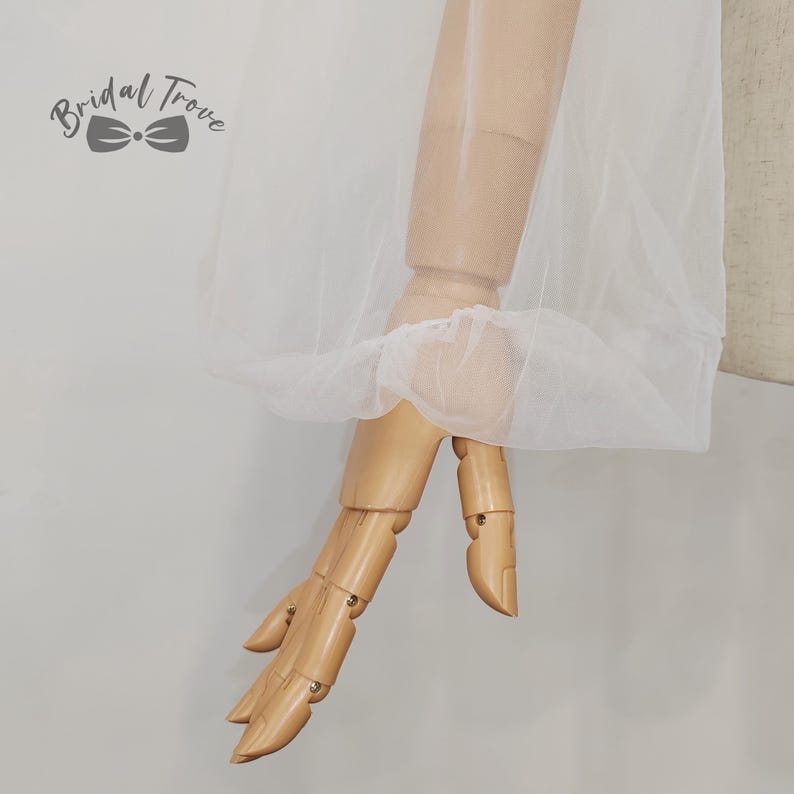 K&ouml;nnte beinhalten: Ein wei&szlig;er T&uuml;llstoff ist &uuml;ber einen h&ouml;lzernen Mannequinarm drapiert. Der Stoff ist am Handgelenk gerafft, wodurch ein R&uuml;scheneffekt entsteht. Das Bridal Trove-Logo ist in der oberen linken Ecke sichtbar. Der Hintergrund ist neutral.