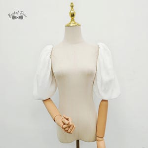 Puede incluir: Una blusa blanca con mangas abullonadas exhibida en un maniquí. Las mangas son voluminosas y fruncidas en el hombro y la muñeca. El maniquí es de color beige neutro y el fondo es blanco liso. El logotipo de Bridal Trove es visible en la esquina superior izquierda.