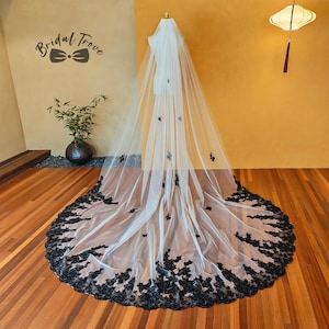 May include: A long, white bridal veil with black floral lace trim and scattered black butterfly accents. The veil is displayed on a mannequin, showcasing its flowing design. The text "Bridal Trove" is visible in the upper left corner.