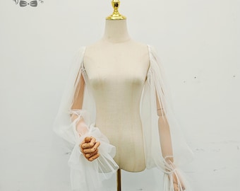 May include: A white, sheer bridal top with long, flowing sleeves. The sleeves are made of a delicate, airy fabric, and the cuffs are gathered with ruffles. The top is displayed on a mannequin with a gold-colored top.