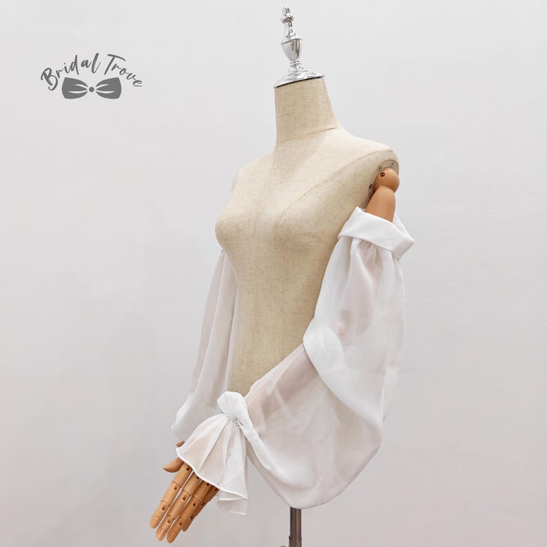 May include: A white, sheer bridal sleeve with a gathered, puffy design and a cuff with a bow detail. The sleeve is displayed on a mannequin. The Bridal Trove logo is in the upper left corner.