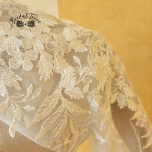 May include: Close-up of a cream-colored bridal garment with intricate floral and leaf embroidery. The delicate lace overlay features a detailed pattern of flowers and leaves. The text "Bridal Trove" is visible.