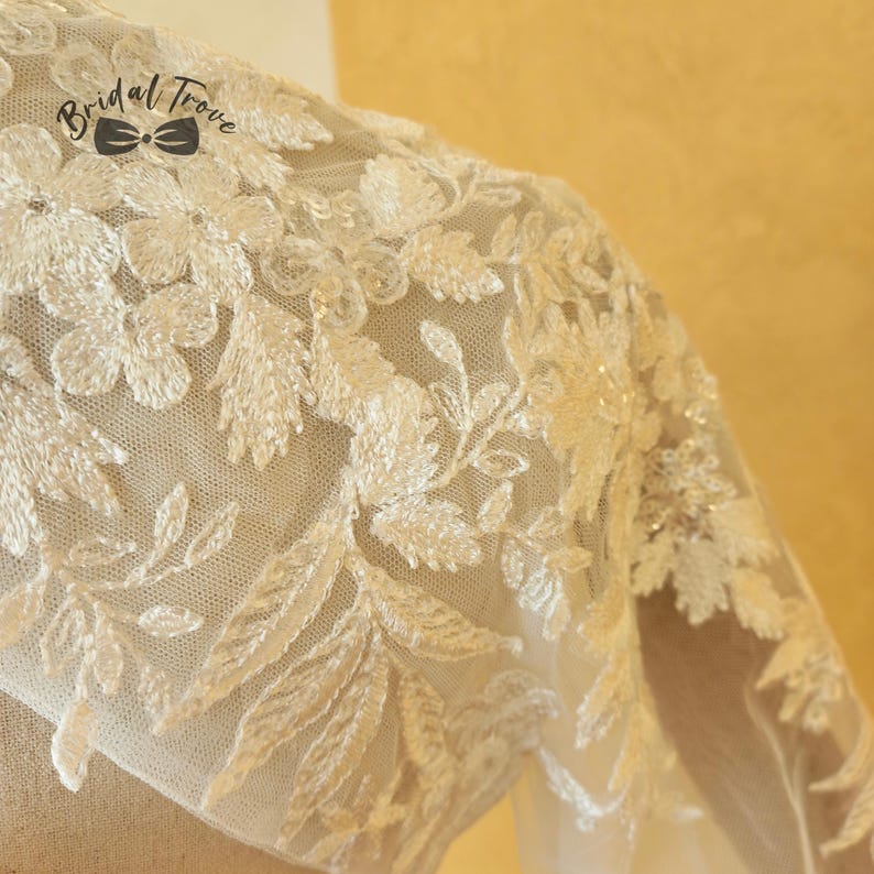 May include: Close-up of a cream-colored bridal garment with intricate floral and leaf embroidery. The delicate lace overlay features a detailed pattern of flowers and leaves. The text "Bridal Trove" is visible.