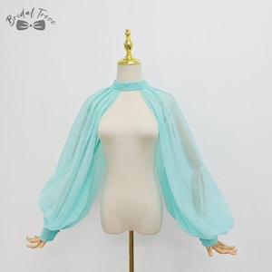 May include: A light blue sheer bridal bolero with long, voluminous sleeves. The bolero features a high neckline with a keyhole opening at the chest. The garment is displayed on a mannequin with a gold-colored top.