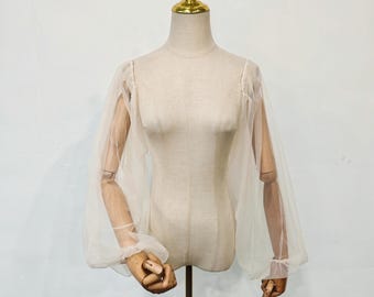 May include: A sheer, off-white blouse with voluminous, long sleeves. The sleeves gather at the wrists with a delicate cuff. The garment is displayed on a mannequin with wooden arms and a golden neck. The background is a plain white wall.