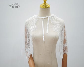 Może przedstawiać: Biała koronkowa peleryna ślubna z delikatnym kwiatowym wzorem. Peleryna ma krótkie rękawy i wiązanie przy szyi. Logo Bridal Trove jest widoczne w lewym górnym rogu.