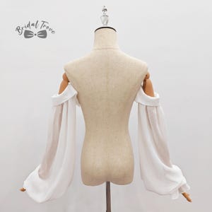 May include: A white, off-the-shoulder bridal top with long, sheer sleeves. The sleeves are gathered at the wrist, creating a flowing, elegant look. The top is displayed on a mannequin, showcasing the design and fit. The logo "Bridal Trove" is visible.
