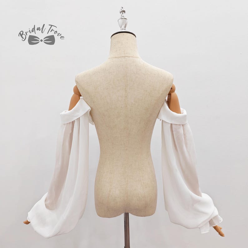 May include: A white, off-the-shoulder bridal top with long, sheer sleeves. The sleeves are gathered at the wrist, creating a flowing, elegant look. The top is displayed on a mannequin, showcasing the design and fit. The logo "Bridal Trove" is visible.