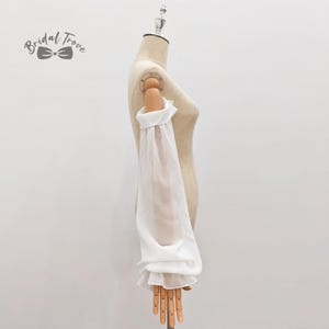 May include: A white, sheer sleeve with a ruffled cuff and shoulder detail is displayed on a mannequin. The sleeve is long and billowy, made of a lightweight fabric. The Bridal Trove logo is in the upper left corner.