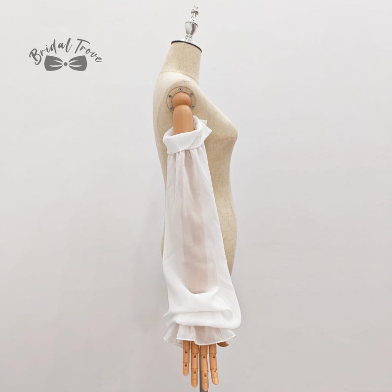 May include: A white, sheer sleeve with a ruffled cuff and shoulder detail is displayed on a mannequin. The sleeve is long and billowy, made of a lightweight fabric. The Bridal Trove logo is in the upper left corner.