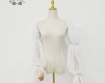 May include: White, sheer, long-sleeved bridal top with voluminous, gathered sleeves. The sleeves are attached to a mannequin. The top is displayed on a white background. The Bridal Trove logo is in the upper left corner.