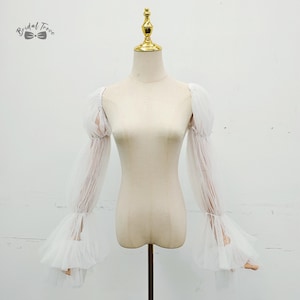 Puede incluir: Mangas de tul blanco con puños con volantes y detalles fruncidos, exhibidas en un maniquí. Las mangas tienen un diseño romántico y fluido, perfecto para novias o eventos formales. El maniquí tiene una parte superior dorada.