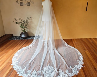 May include: A long, white bridal veil with intricate lace detailing along the bottom edge. The veil is displayed on a mannequin, showcasing its flowing design. The text "Bridal Trove" is visible in the upper left corner.