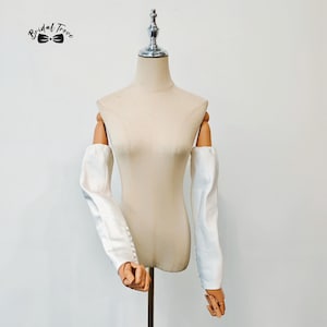 May include: A white satin bridal sleeve set on a mannequin. The sleeves are off-the-shoulder with buttoned cuffs. The text "Bridal Trove" is in the upper left corner. The mannequin is on a wooden stand.