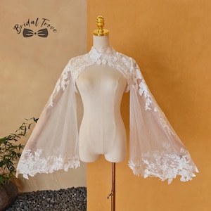 Op de afbeelding: Een witte, doorschijnende bruidscape met bloemenkanten details. De cape heeft een hoge halslijn en uitlopende mouwen. Het Bridal Trove-logo is zichtbaar. De cape wordt op een mannequin gepresenteerd.