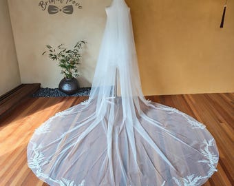 May include: A long, white bridal veil with a detailed, embroidered floral design along the edge. The veil is displayed on a mannequin, showcasing its length and delicate fabric. The background includes a wooden floor and a wall with the text "Bridal Trove".
