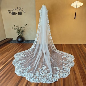 May include: A long, white bridal veil with delicate floral embellishments. The veil is displayed on a mannequin, showcasing its intricate design and flowing fabric. The flowers are clustered along the edges and cascade down the veil.