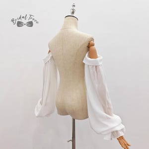 May include: A white off-the-shoulder top with long, flowing sleeves. The sleeves are gathered at the wrist and have a slight puff. The top is displayed on a mannequin. The text "Bridal Trove" is in the upper left corner.