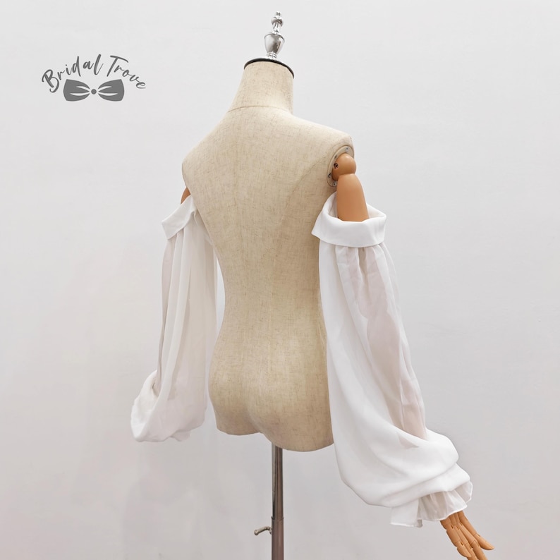 May include: A white off-the-shoulder top with long, flowing sleeves. The sleeves are gathered at the wrist and have a slight puff. The top is displayed on a mannequin. The text "Bridal Trove" is in the upper left corner.