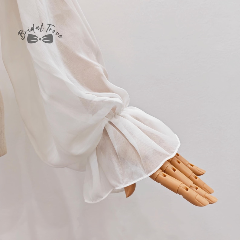 May include: Close-up of a white, sheer sleeve with a ruffled cuff. The sleeve is gathered at the wrist, creating a voluminous effect. The fabric appears delicate and flowing. The logo "Bridal Trove" is visible in the upper left corner.