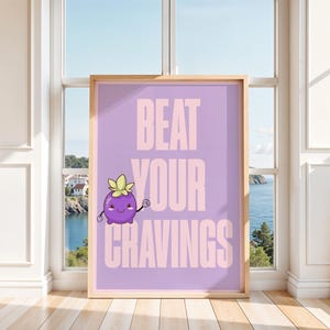 May include: A framed poster with the text "BEAT YOUR CRAVINGS" in light pink on a lavender background. A cartoon purple fruit with a green leafy top is featured. The poster is displayed in front of a window overlooking a coastal scene.