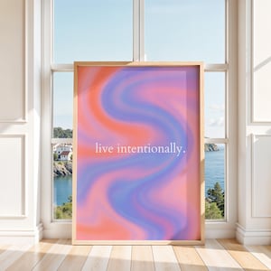 May include: A framed art print featuring an abstract design in orange, pink, and blue hues. The white text reads "live intentionally." The artwork is displayed in a room with a window overlooking a coastal scene, with a wooden frame.