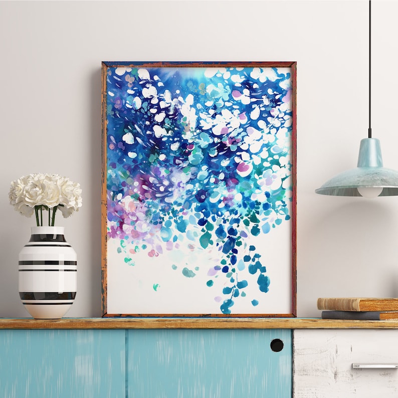 May include: Abstract watercolor art print in a wooden frame. The artwork features a cascade of blue, teal, purple, and white brushstrokes. The framed art is displayed on a blue cabinet with a vase of white flowers and a striped vase.