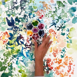 May include: A watercolor painting of a floral pattern with a hand holding a round watercolor palette with six paint wells. The palette is filled with different colors of paint, including blue, green, red, and yellow.