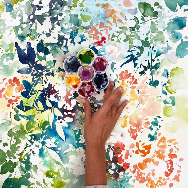 May include: A watercolor painting of a floral pattern with a hand holding a round watercolor palette with six paint wells. The palette is filled with different colors of paint, including blue, green, red, and yellow.