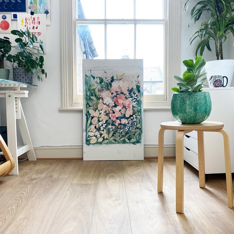 Op de afbeelding: Een abstract bloemenschilderij met roze en witte bloemen en groen blad. Het kunstwerk wordt tentoongesteld in een lichte kamer met een raam, naast een kleine houten kruk met een groene potplant.