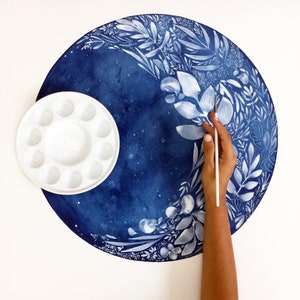 Puede incluir: Una paleta de pintura blanca con 12 huecos se encuentra sobre una pintura de acuarela azul y blanca con un dise&ntilde;o floral. Una mano sostiene un pincel blanco sobre la pintura.