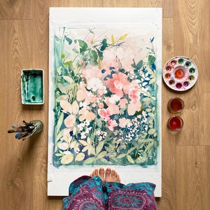 Op de afbeelding: Aquarel schilderij van roze en witte bloemen met groen blad. Een palet met aquarelverf, penselen en verf zijn rond het kunstwerk gerangschikt. De schildering staat op een wit oppervlak en de achtergrond is een houten vloer.