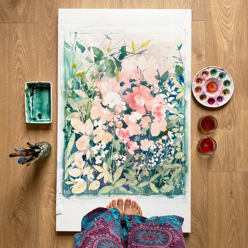 Op de afbeelding: Aquarel schilderij van roze en witte bloemen met groen blad. Een palet met aquarelverf, penselen en verf zijn rond het kunstwerk gerangschikt. De schildering staat op een wit oppervlak en de achtergrond is een houten vloer.