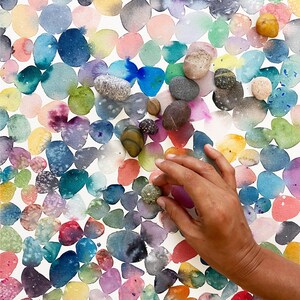 以下が含まれることがあります： 手は、小さな質感のある緑色の石をそっと触れています。石は、カラフルで重なり合う円の水彩画の上にあります。円はピンク、青、黄色、紫の色合いで、いくつかの天然石が散らばっています。