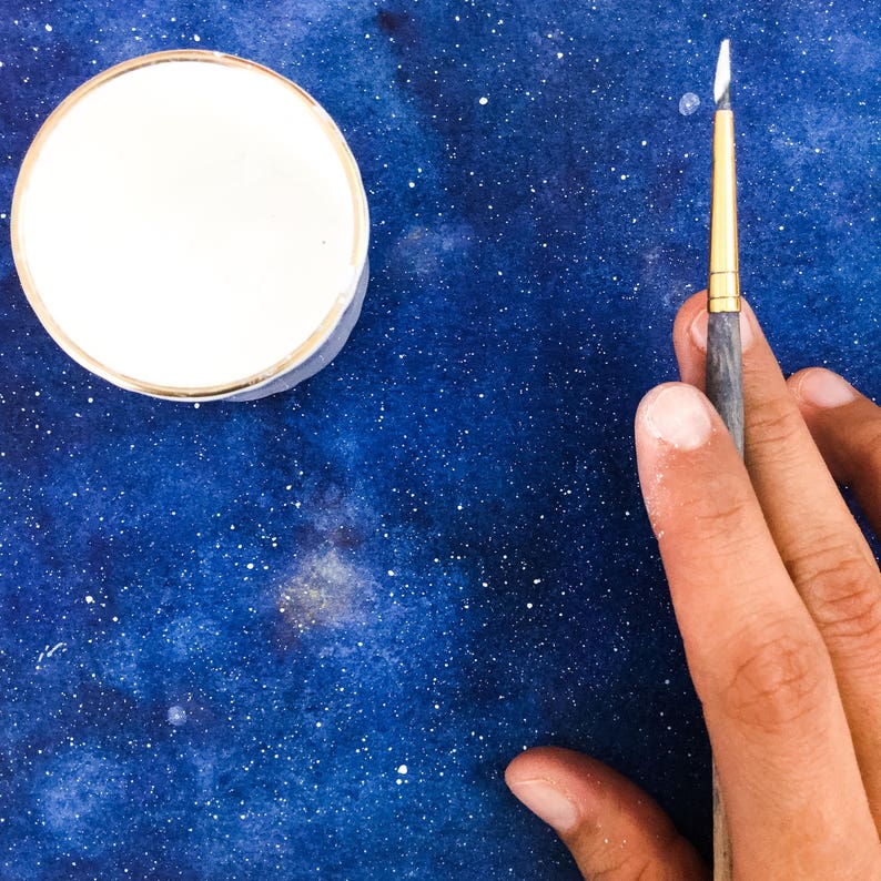 May include: A close-up of a hand holding a paintbrush over a blue watercolor background with white dots. A white paint container is in the upper left corner of the image.