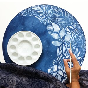 Puede incluir: Una paleta de pintura de cer&aacute;mica blanca con 12 pozos, una pintura de acuarela azul de dise&ntilde;os florales blancos sobre un lienzo redondo y una mano sosteniendo un pincel plateado.