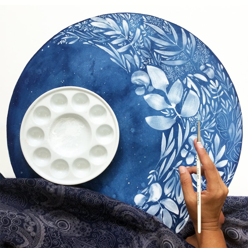 Puede incluir: Una paleta de pintura de cer&aacute;mica blanca con 12 pozos, una pintura de acuarela azul de dise&ntilde;os florales blancos sobre un lienzo redondo y una mano sosteniendo un pincel plateado.