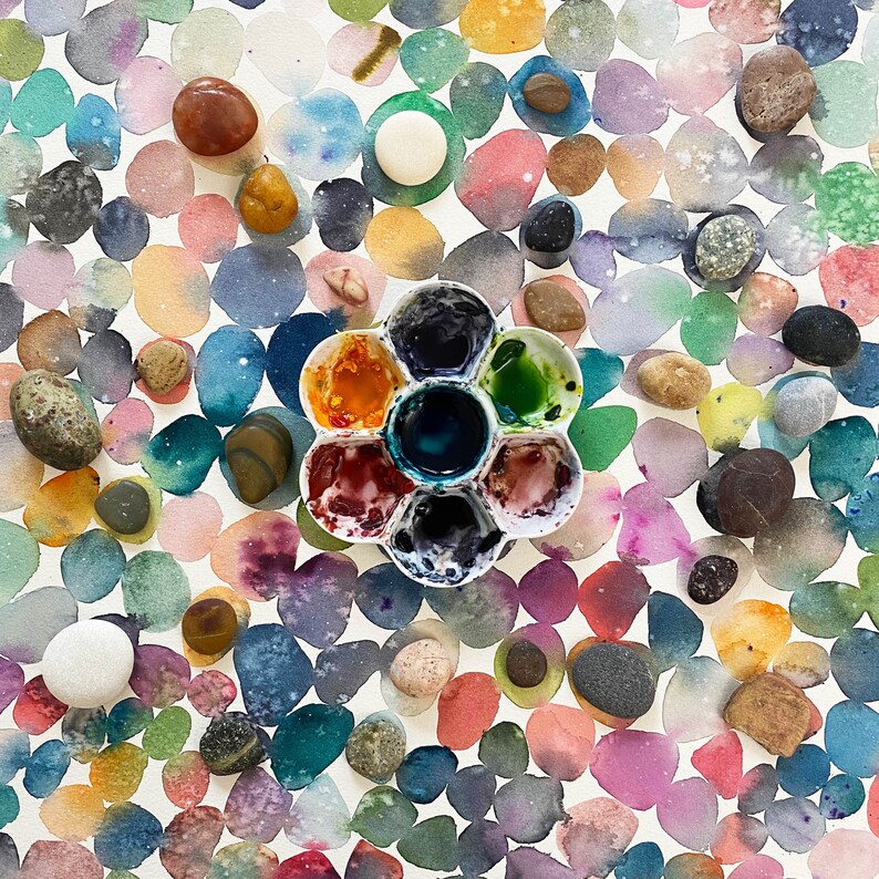 以下が含まれることがあります： パステルカラーの重なり合う円のパターンが特徴的な水彩画。さまざまな色と質感の小さな滑らかな小石が表面に散らばっています。さまざまな色で満たされた花型のペイントパレットが中央にあります。