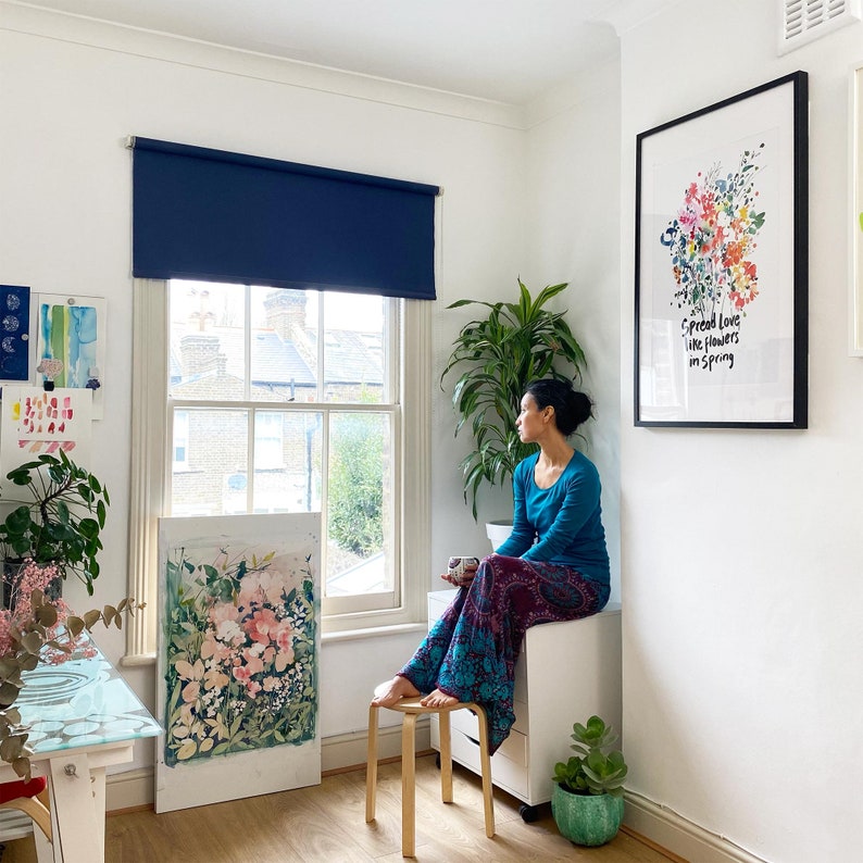 Op de afbeelding: Een vrouw zit bij een raam en kijkt naar buiten. Een grote ingelijste print met kleurrijke bloemen en de tekst "Spread love like flowers in Spring" hangt aan de muur. Een schilderij van bloemen leunt tegen de muur. Boven het raam hangt een donkerblauw rolgordijn.