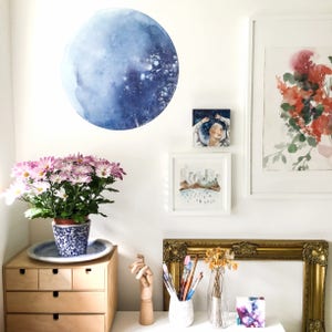 May include: A white wall with a blue watercolour moon, a framed watercolour painting of a city skyline, a framed watercolour painting of a woman with her arms raised, and a framed watercolour painting of a floral arrangement. There is a potted plant with pink flowers in the foreground, a wooden shelf with drawers, and a gold-framed mirror with dried flowers in a vase.