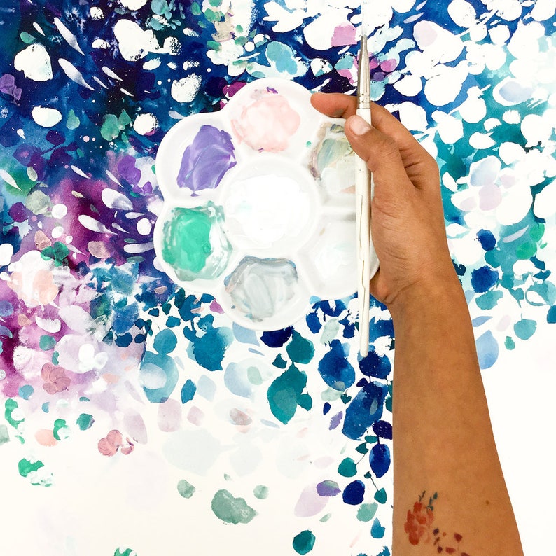 May include: A white plastic paint palette with various colors of paint, including purple, green, and pink, is held in front of a colorful abstract painting. A paintbrush is held in the hand. The background features a vibrant watercolor design with blue, purple, and white hues.
