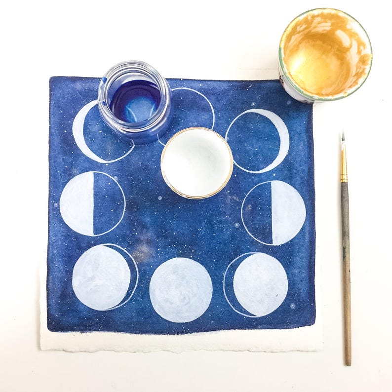 May include: A blue watercolor painting of the moon phases with white paint. The painting is surrounded by a white border. There are two small bowls, one with blue liquid and one with white liquid, and a paintbrush on a white surface.