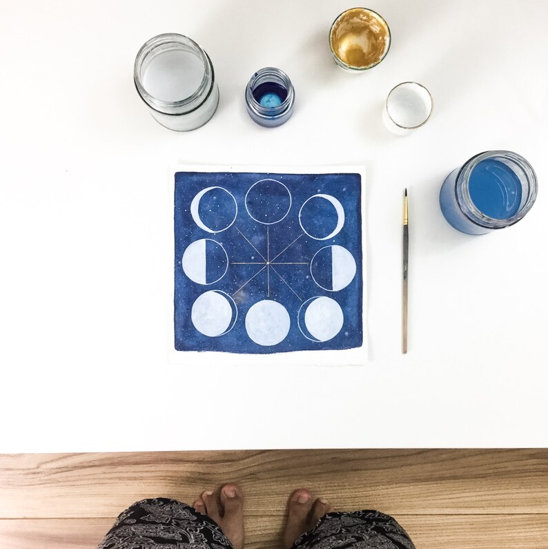 May include: A watercolor painting of the moon phases in a blue starry sky. The painting is surrounded by jars of paint and a paintbrush.