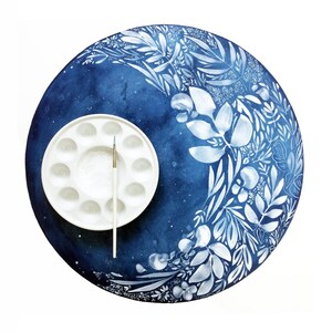 Puede incluir: Una paleta de pintura blanca con 12 huecos y un pincel sobre un lienzo redondo azul y blanco con un dise&ntilde;o floral.