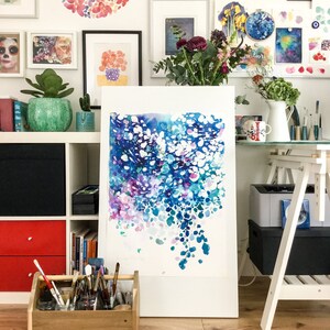 May include: An abstract watercolor painting in shades of blue, purple, and white, displayed on an easel. The artwork features a flowing, organic design with a variety of brushstrokes. The background includes framed art, plants, and art supplies.