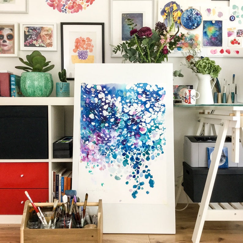 May include: An abstract watercolor painting in shades of blue, purple, and white, displayed on an easel. The artwork features a flowing, organic design with a variety of brushstrokes. The background includes framed art, plants, and art supplies.
