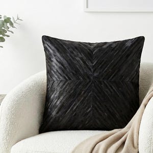 Peut inclure: Un coussin décoratif noir carré avec un motif géométrique de lignes diagonales. Le coussin est posé sur une chaise blanche et incurvée, avec une couverture beige drapée dessus. Un vase blanc avec de la verdure est visible en arrière-plan.