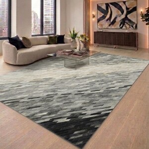May include: A large area rug with a geometric pattern in shades of gray and black dominates the room. The rug's design features a gradient effect, transitioning from light gray to dark gray and black. The rug is placed on a light-colored wooden floor.
